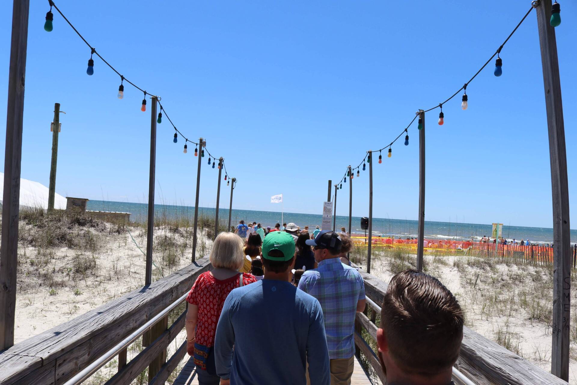 A group of people walks along a wooden boardwalk toward a sandy beach under a clear blue sky, framed by hanging lights.