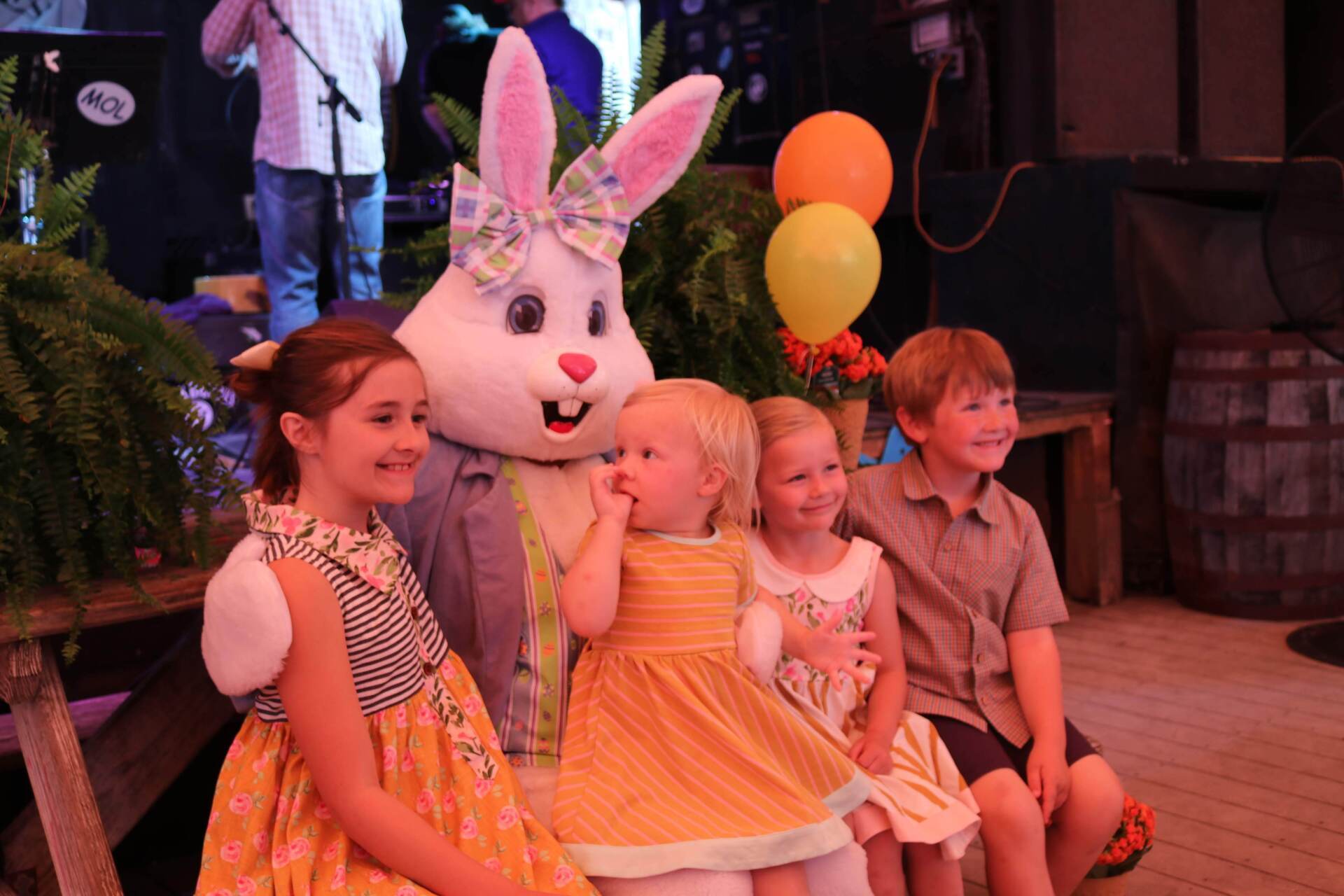 Four children sit with an Easter Bunny mascot outdoors, smiling in front of yellow and orange balloons.