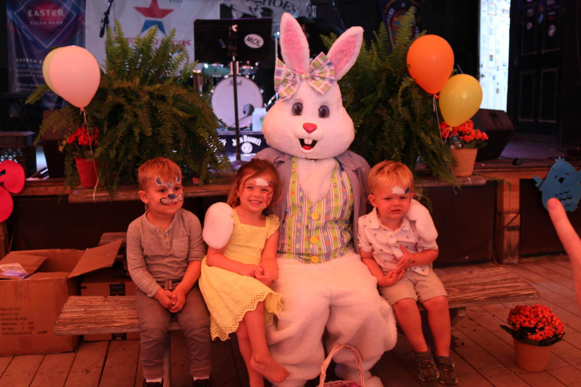 Three children sit with the Easter Bunny on a wooden bench, decorated with balloons and plants in a festive setting.
