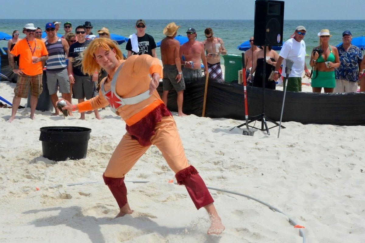 A person dressed as the character He-Man throws a ball on a sandy beach while a crowd of people watches in the background.