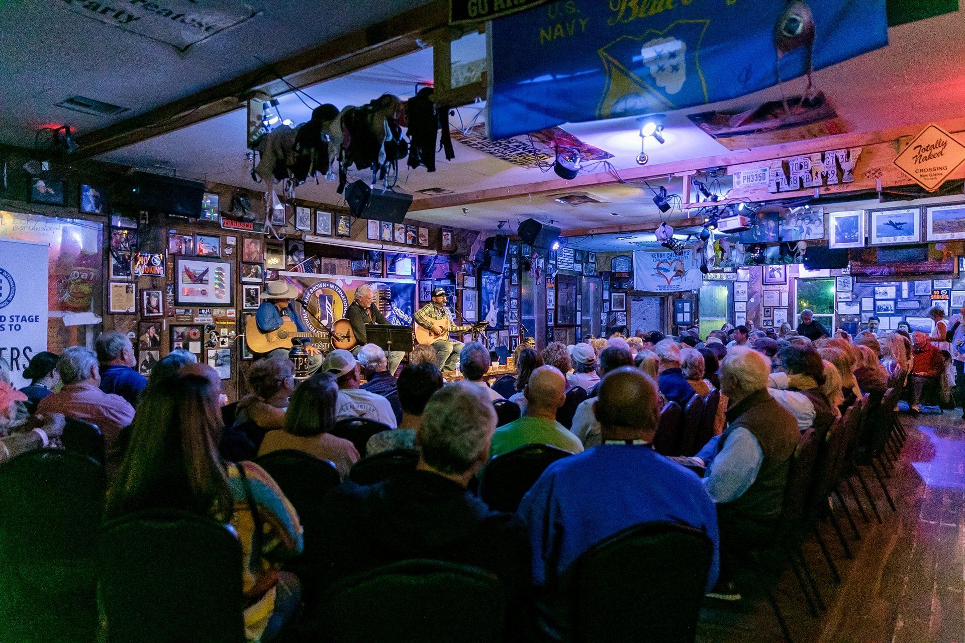 An audience watches a musical performance on a small stage in a crowded, dimly lit venue decorated with banners.