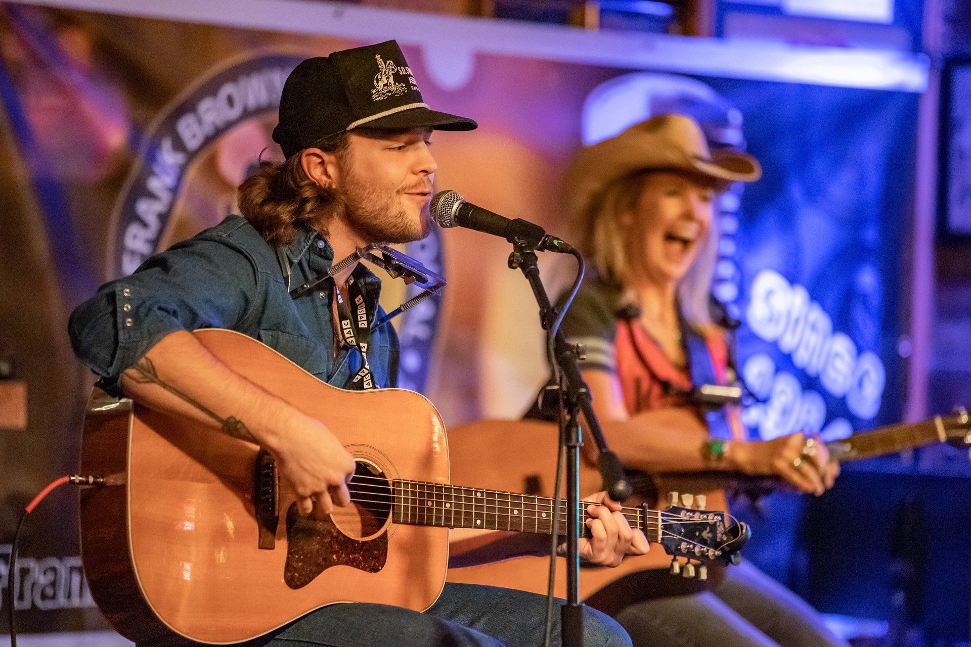 A person in a baseball cap plays acoustic guitar and sings into a microphone, with a smiling partner playing guitar behind.