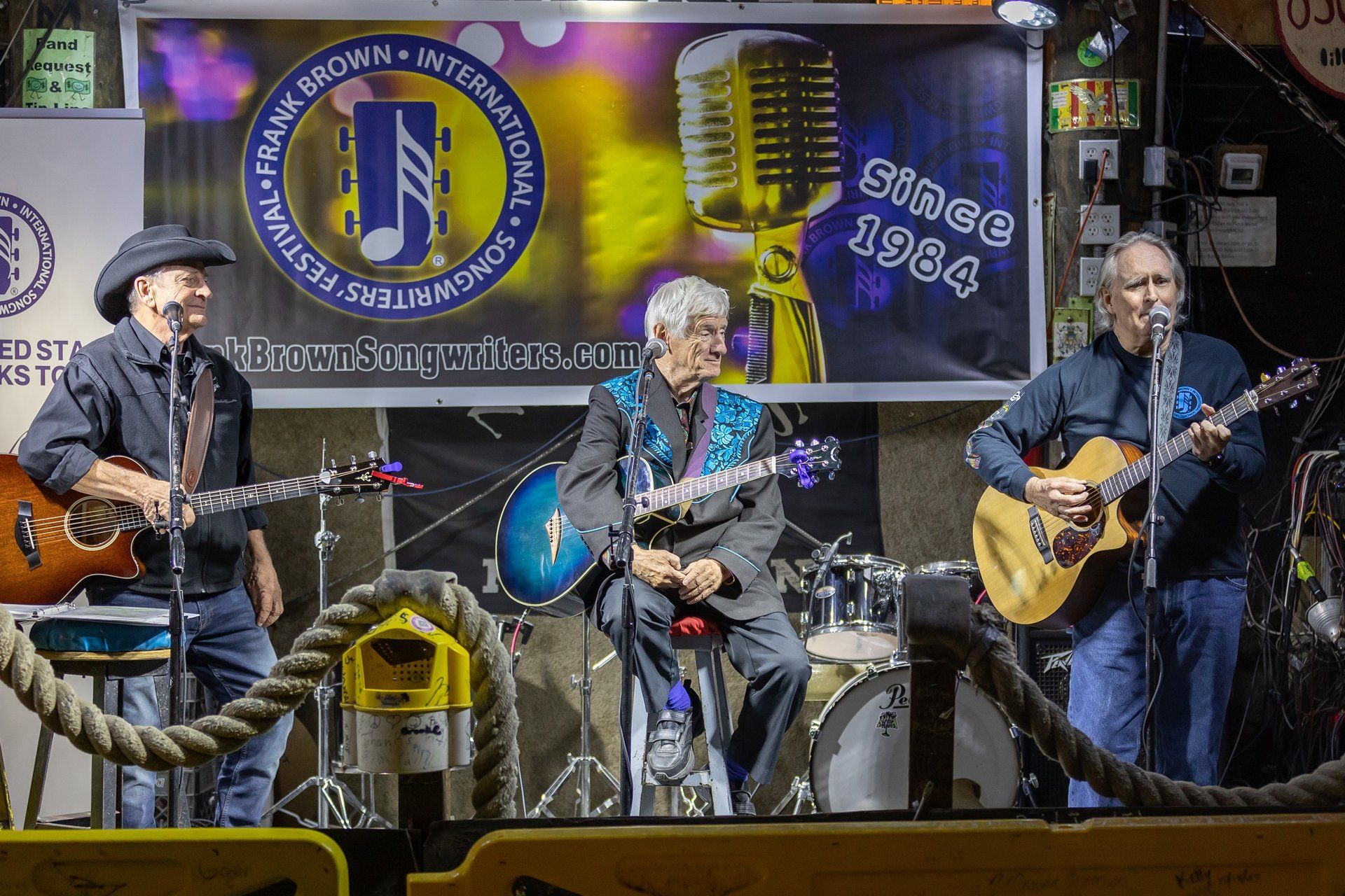 Three musicians play acoustic guitars on stage in front of a banner for the Frank Brown International Songwriters Festival.