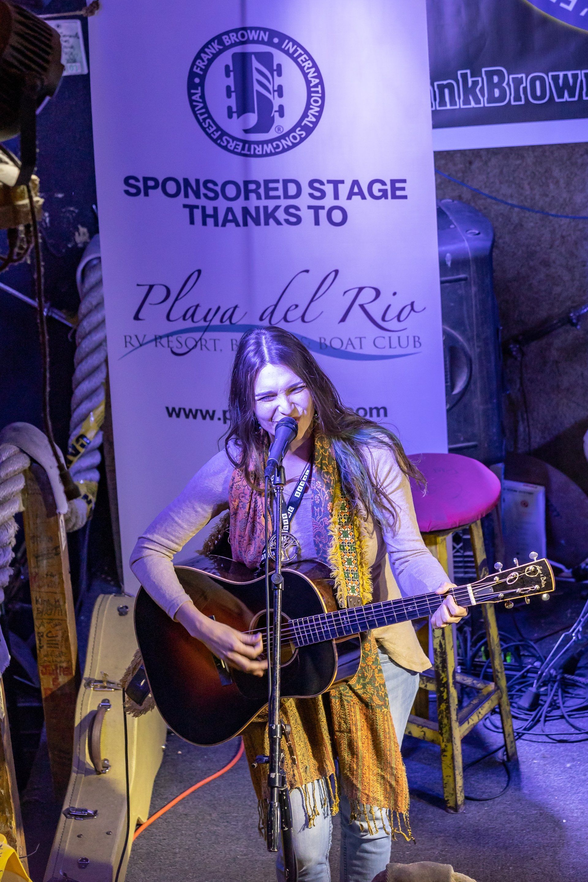 A person playing an acoustic guitar and singing into a microphone on a stage with a banner in the background.