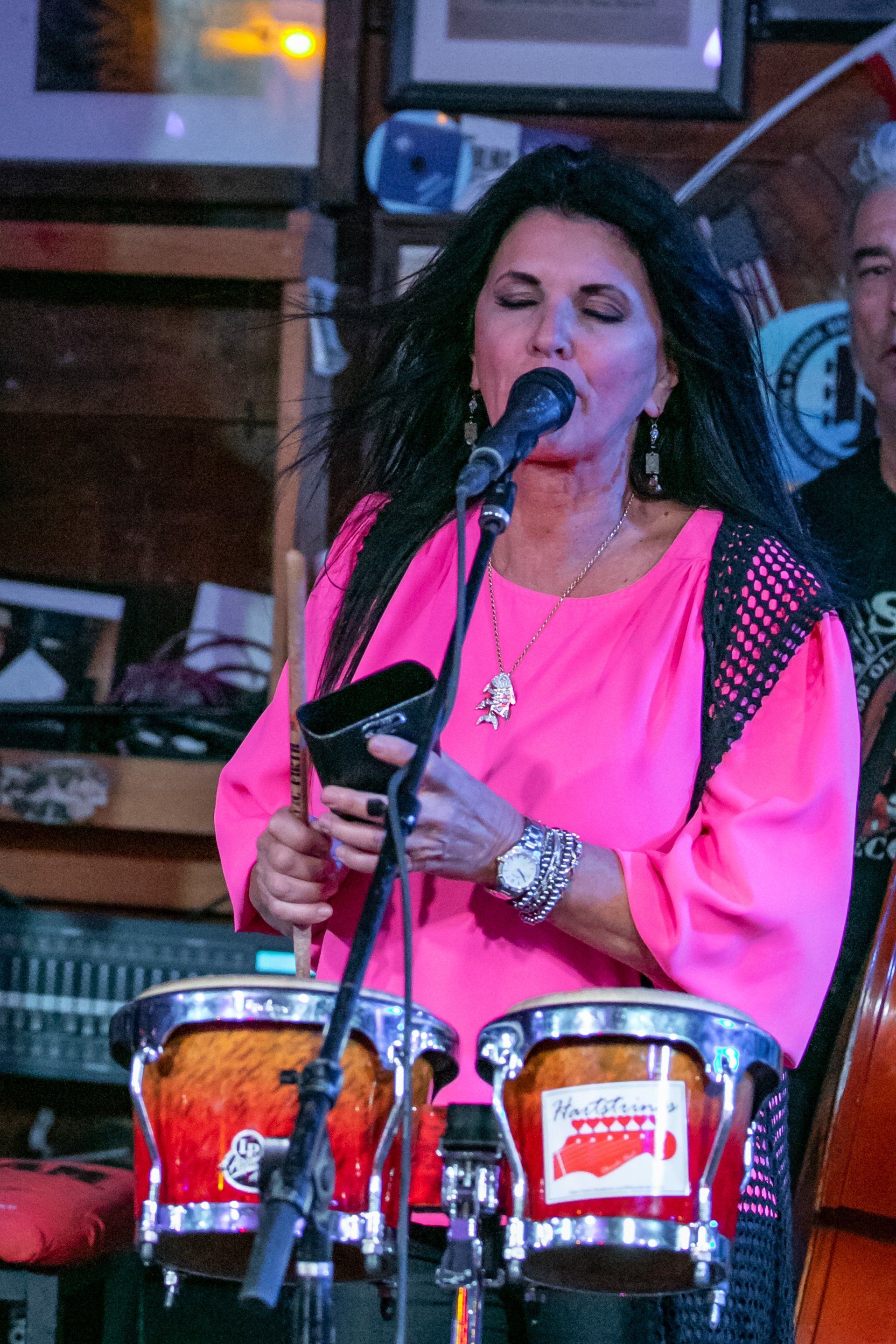 A performer in a bright pink top sings into a microphone while playing a cowbell and bongo drums on stage.