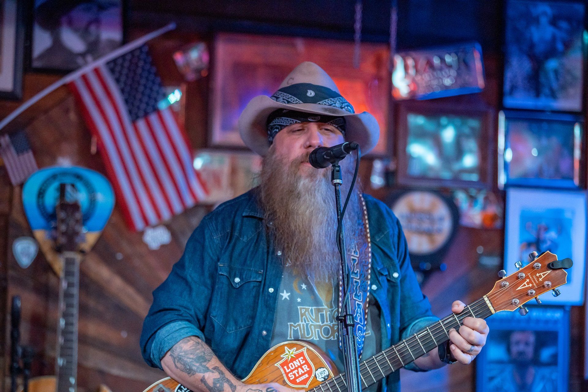 A bearded musician wearing a cowboy hat and denim shirt performs while playing an acoustic guitar in a decorated venue.