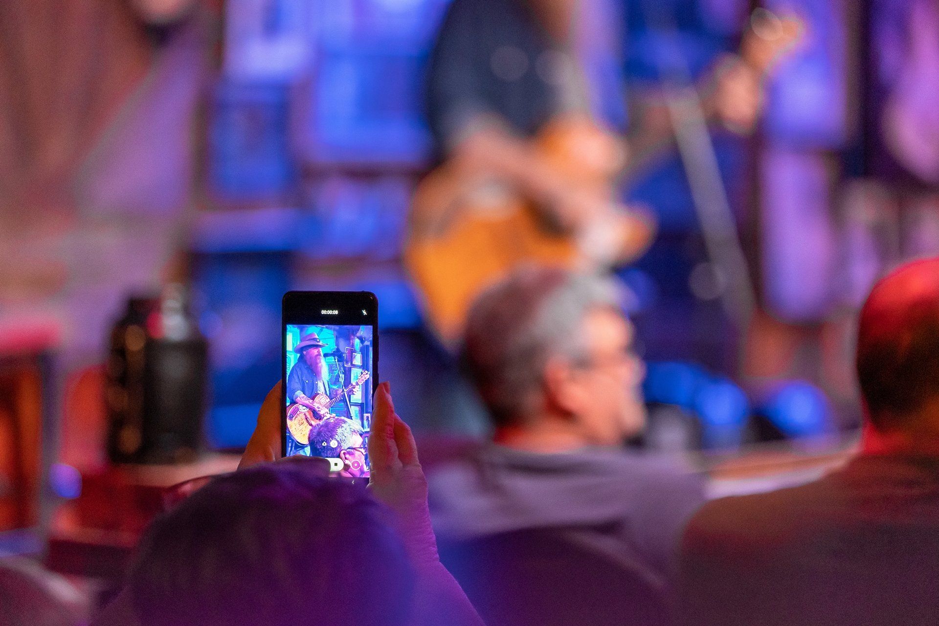 A person holds a phone up to record a concert, capturing a performer on stage in the blurred background.
