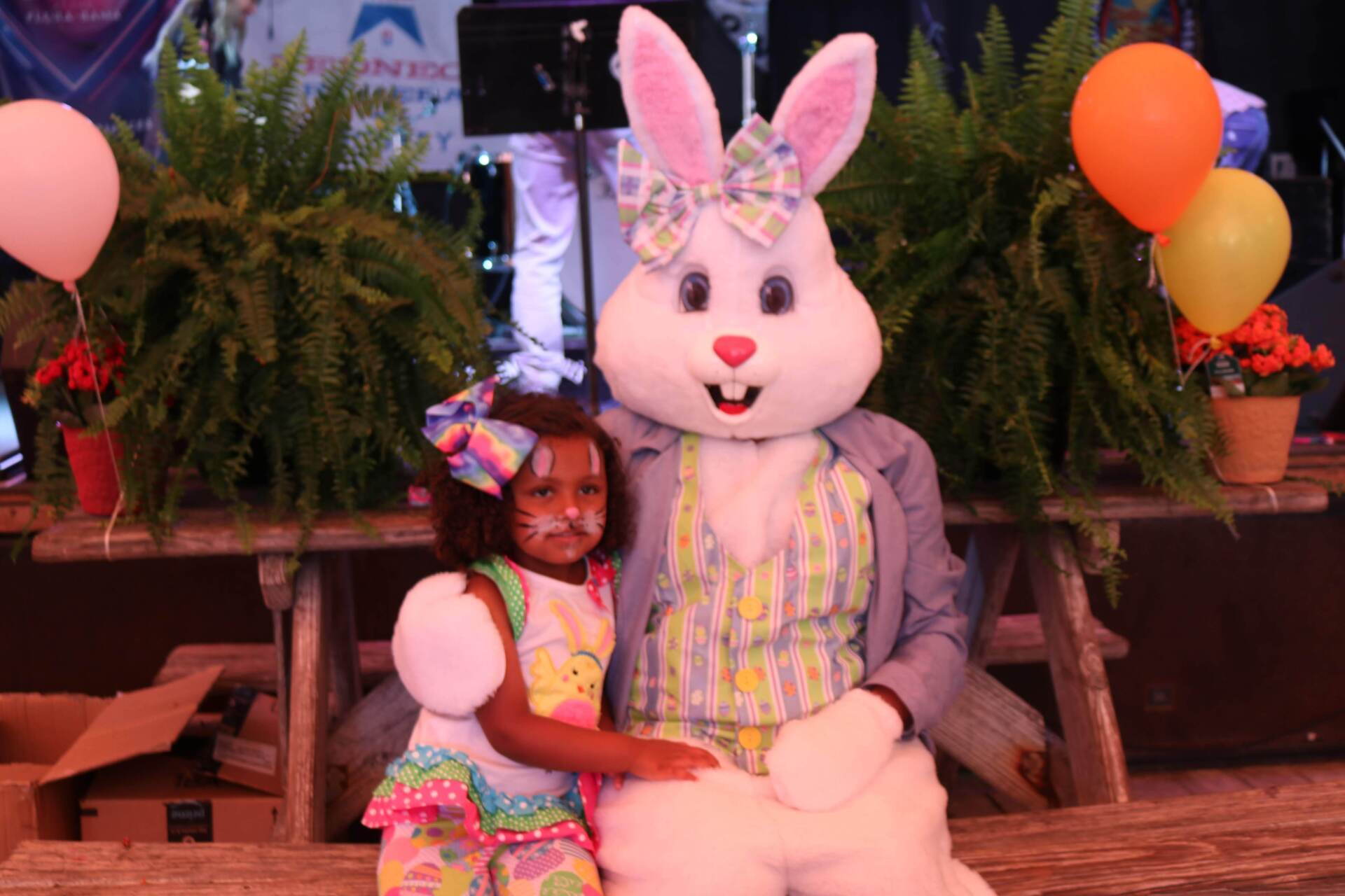 A child in festive clothing poses for a photo with a large Easter Bunny mascot at a party decorated with balloons.