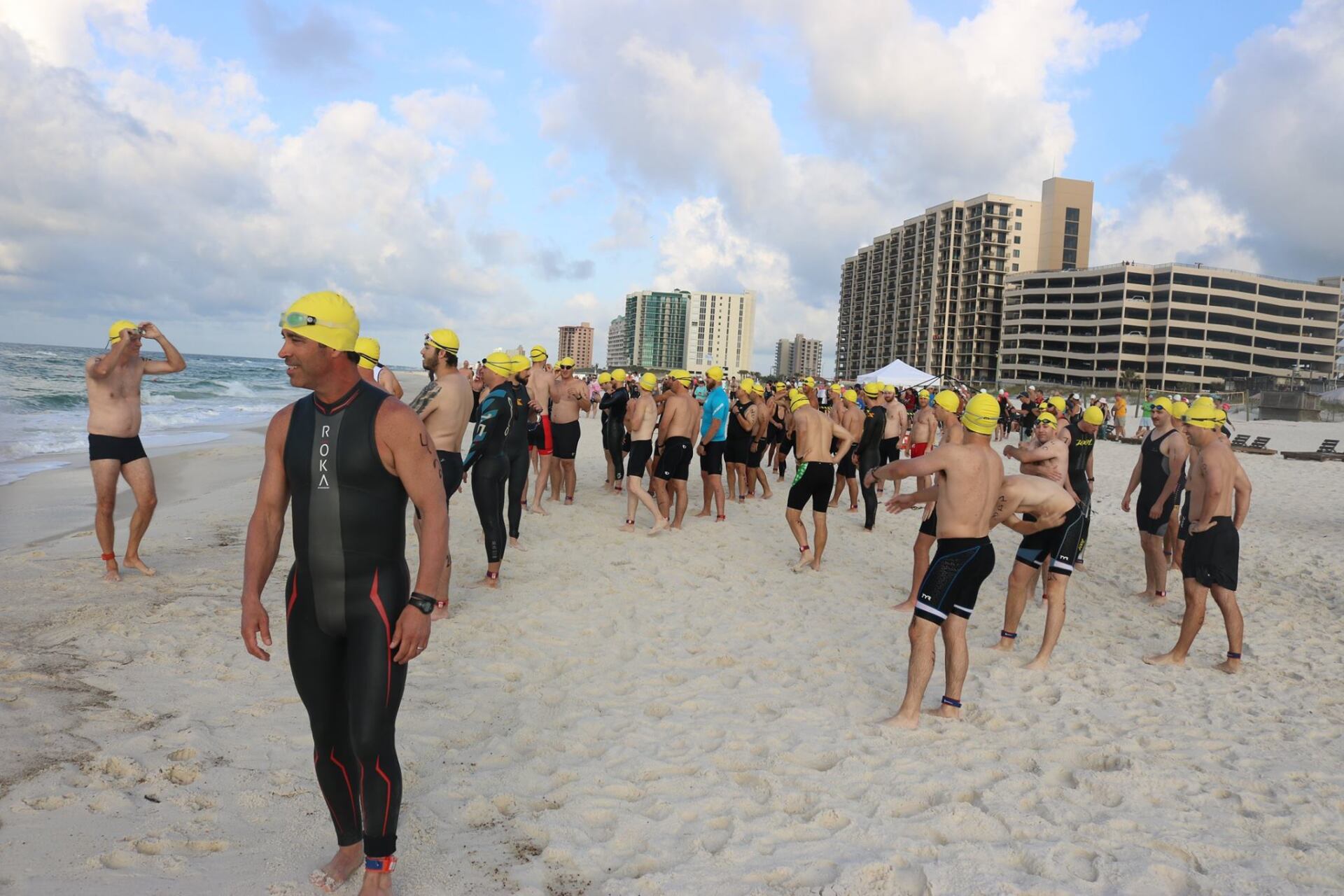 A large group of swimmers wearing yellow caps stands on a sandy beach near high-rise buildings before a race.