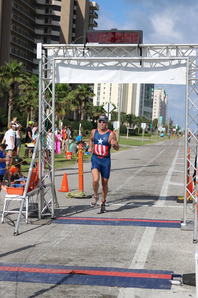 A runner wearing a blue star-spangled jersey runs toward the finish line on a city street near tall buildings.