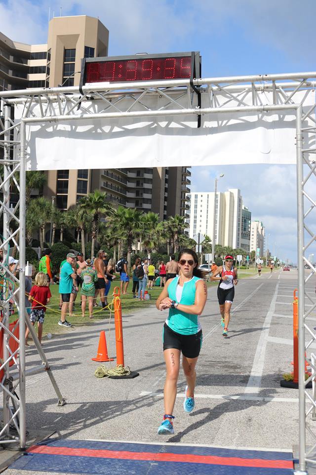 A runner approaches a race finish line with a digital clock overhead, set in a sunny, palm-lined beach area.