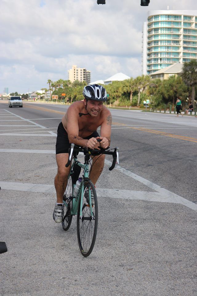 A shirtless cyclist wearing a helmet rides a bike on a paved coastal road near buildings and palm trees.