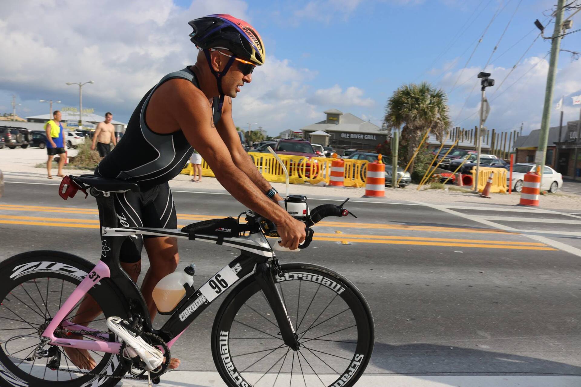 A cyclist in a black and white triathlon suit rides a black and pink racing bike along a paved road near a beach.