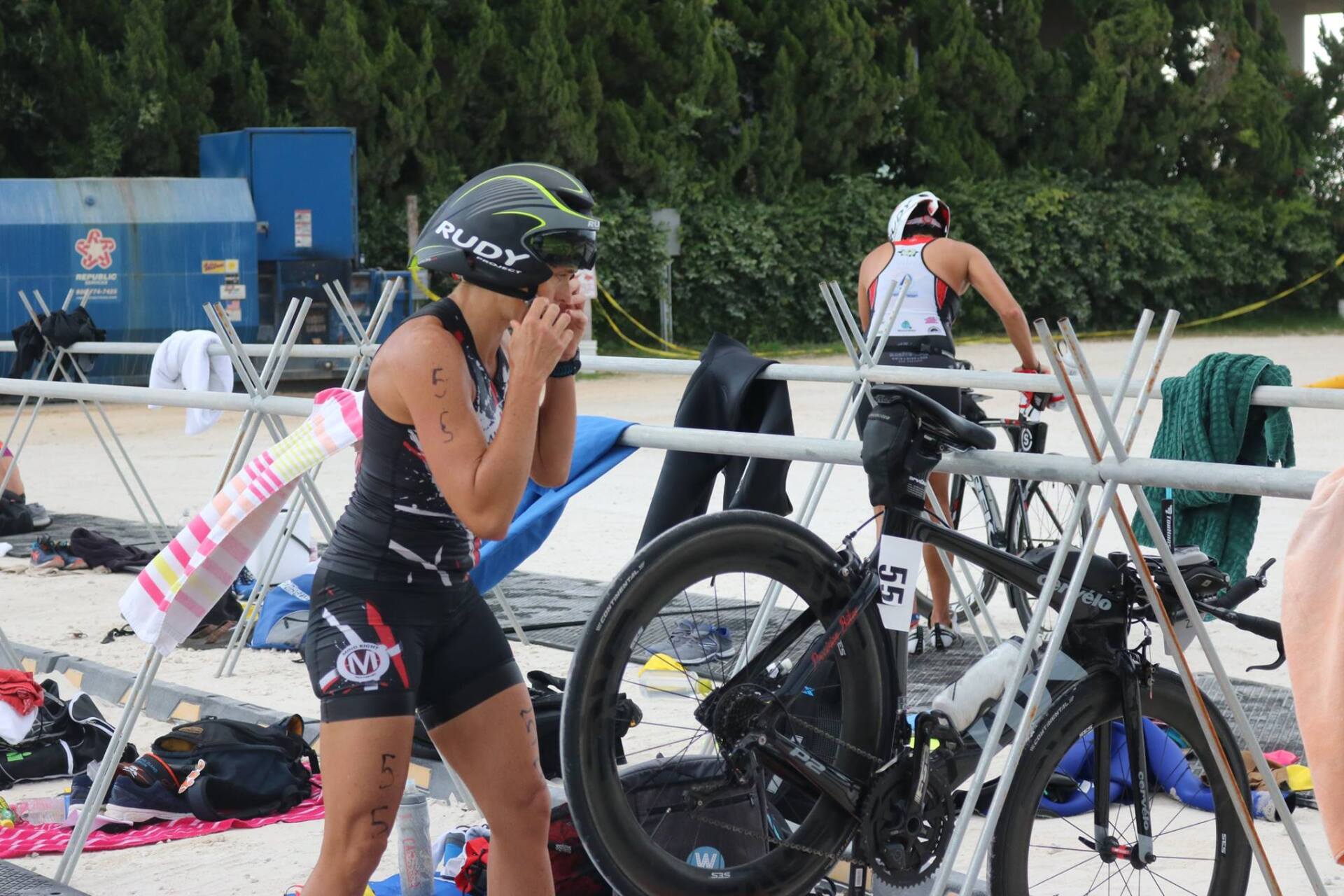 A triathlete in a black kit adjusts their helmet in a transition area beside a bicycle, with another athlete nearby.