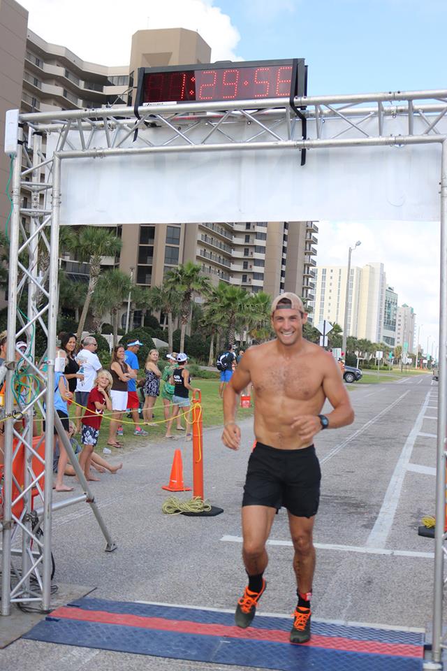 A person running under a race finish line arch with a digital clock displaying 1:24:54 on an outdoor paved road.