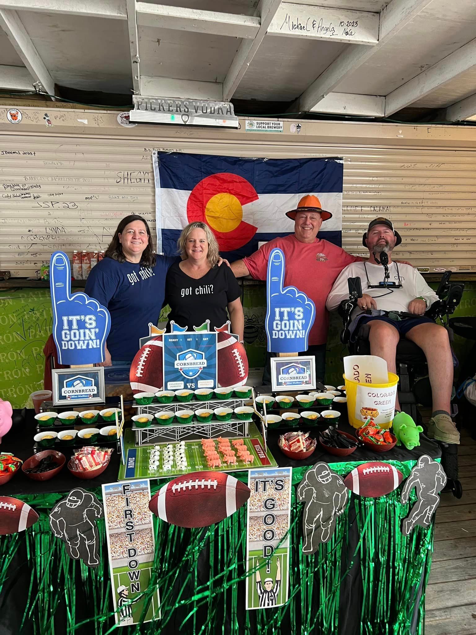 Four people pose behind a table decorated for a football party, with a Colorado flag hanging on the wall behind them.
