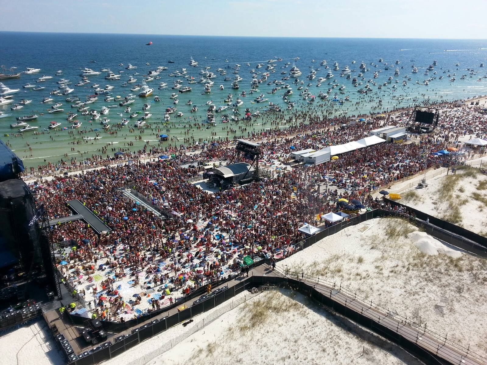 Aerial view of a packed sandy beach music festival with a large crowd and numerous boats anchored in the water nearby.