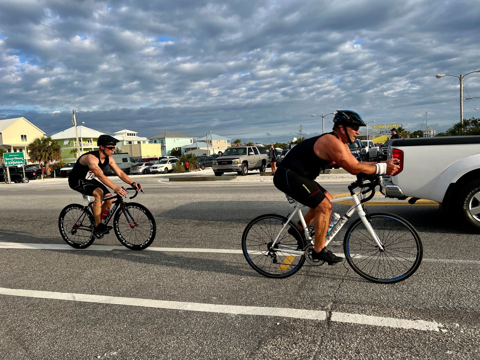 Two cyclists in athletic gear ride their road bikes down a paved street with a pickup truck nearby under a cloudy sky.