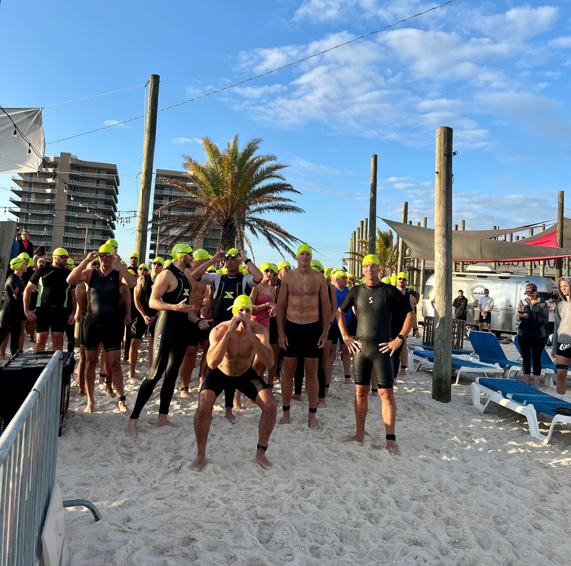 A group of swimmers in yellow caps and wetsuits gather on a sandy beach before an open-water race.