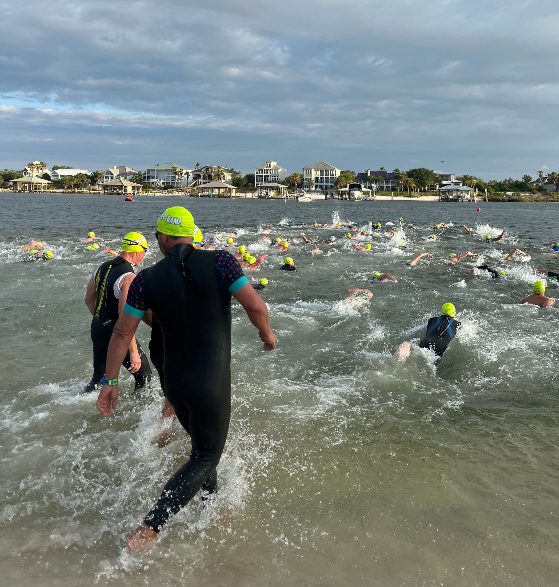 People in wetsuits and bright caps wading and swimming into a body of water during a triathlon start.