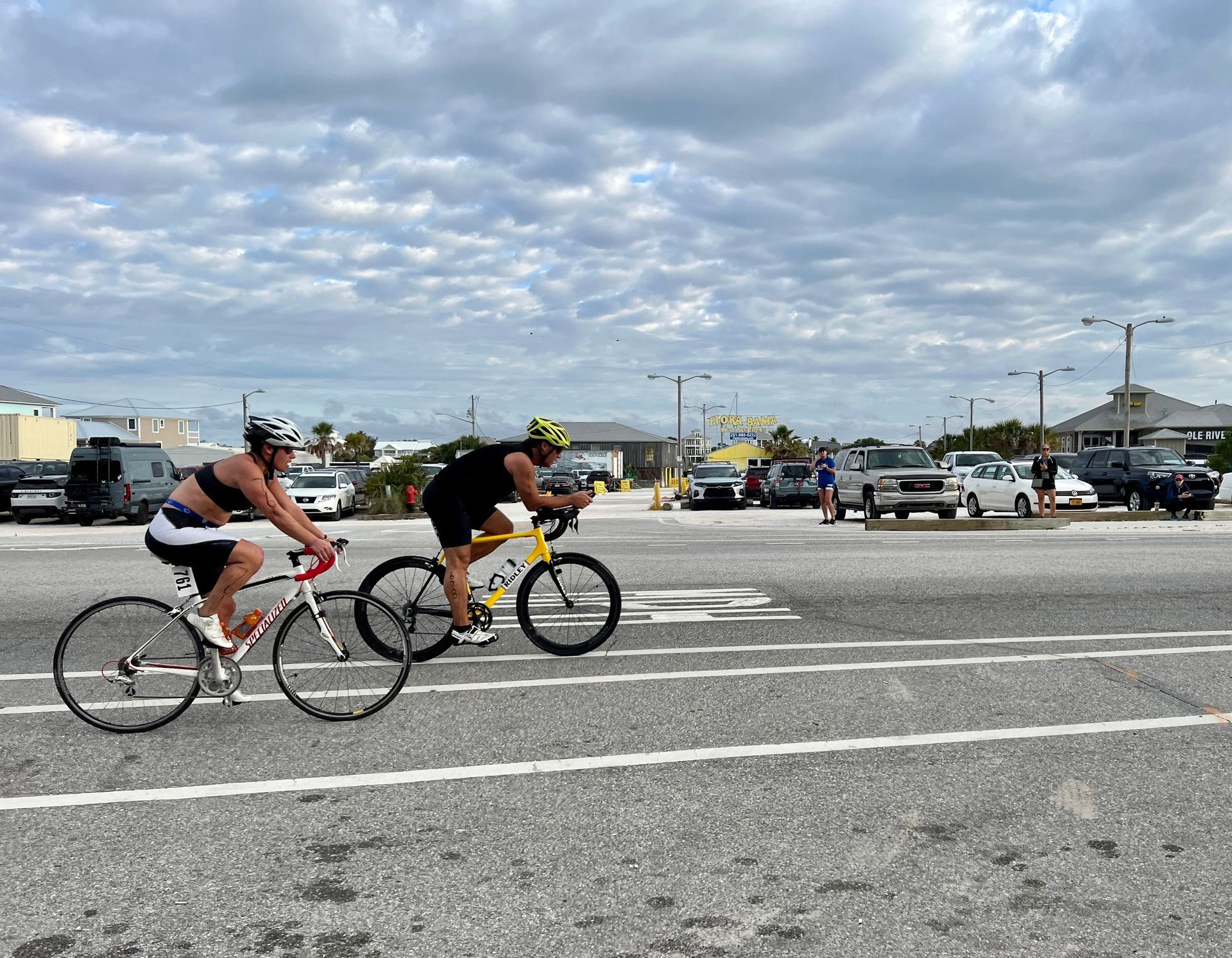 Two cyclists ride side-by-side on a paved road under a cloudy sky with vehicles parked in the background.