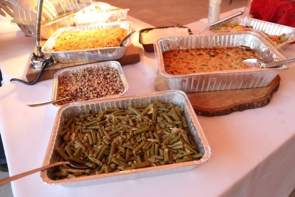 A buffet table featuring aluminum trays filled with green beans, rice, and baked dishes on a white tablecloth.