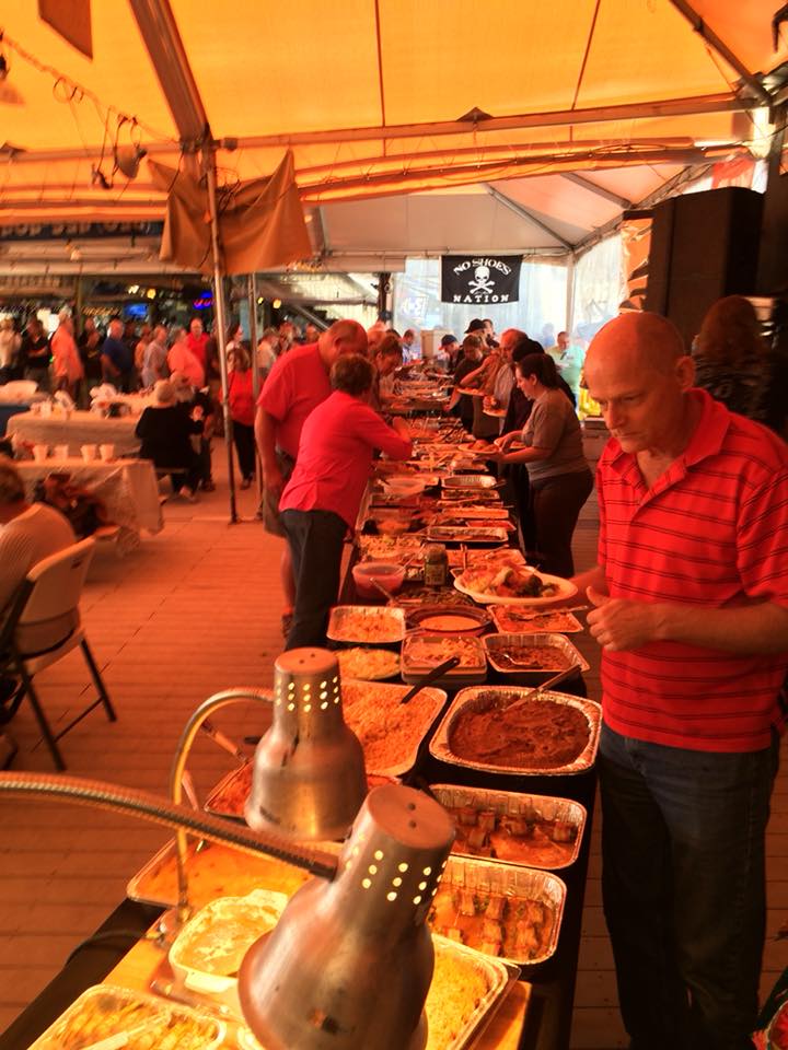 People move through a buffet line under a yellow-lit tent, filling plates from various metal food trays.