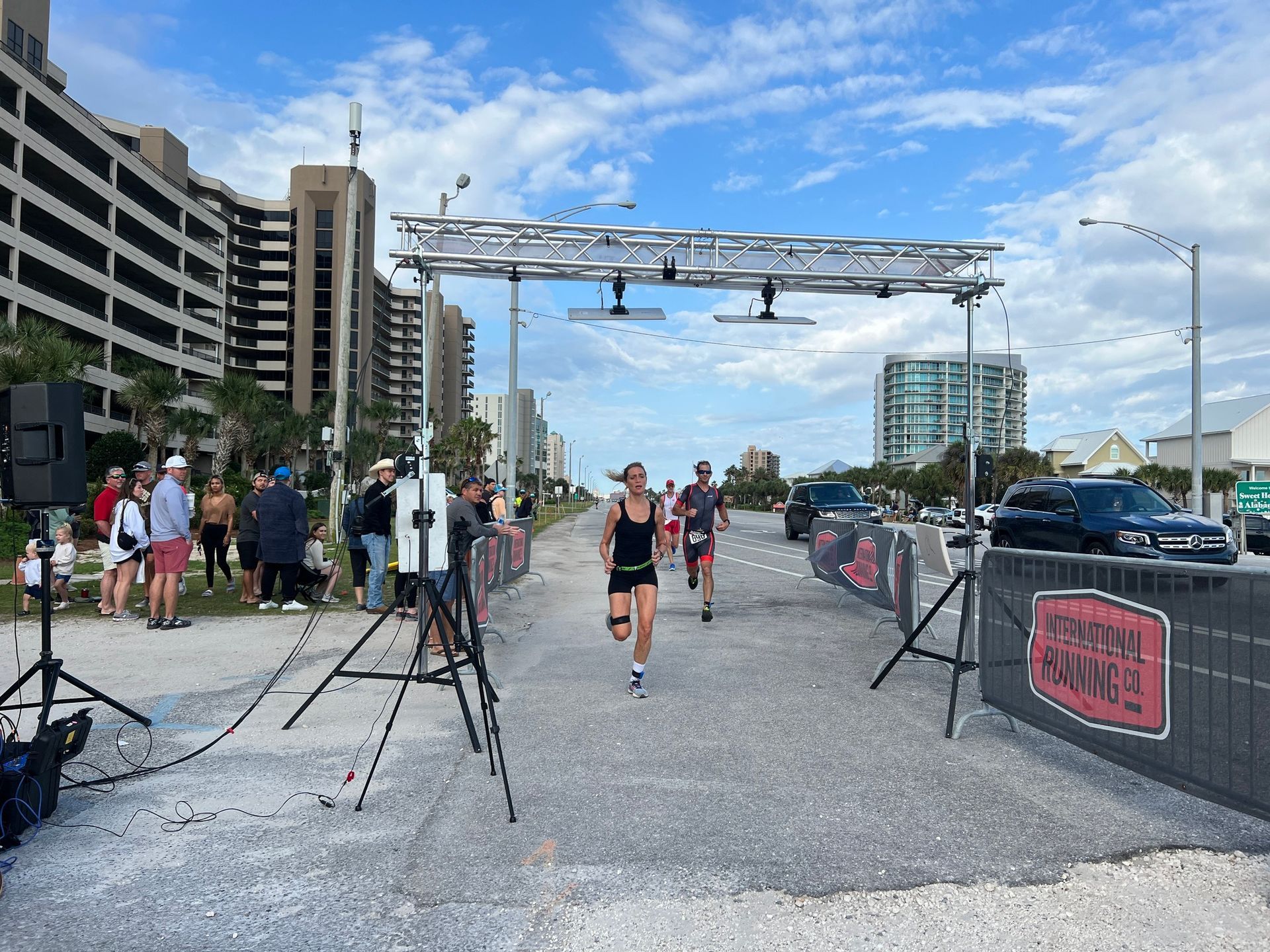 Two people run toward a finish line structure on a paved path near a beach, with onlookers and buildings nearby.