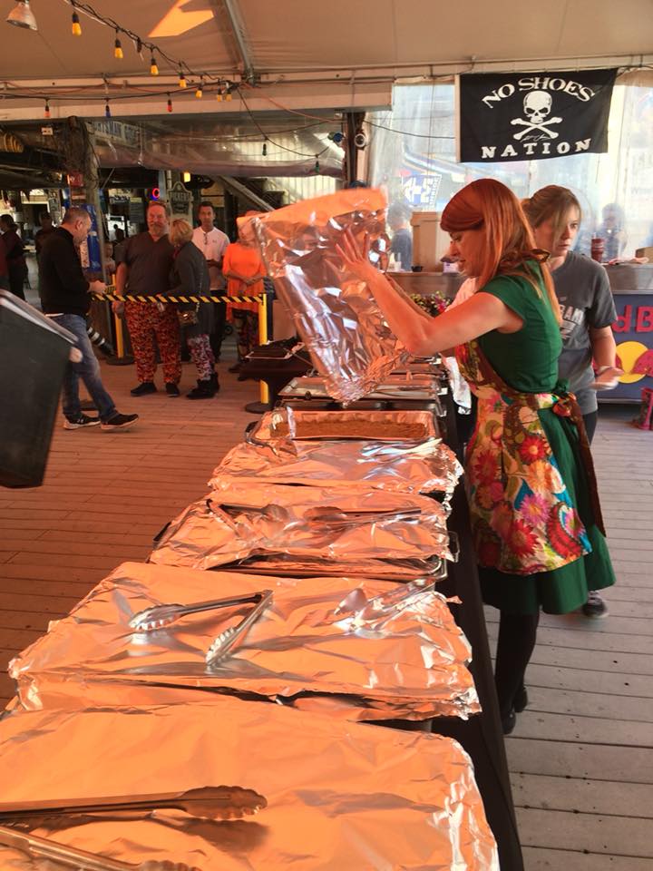 A person wearing a floral apron prepares a buffet table covered in aluminum foil under a tent with a