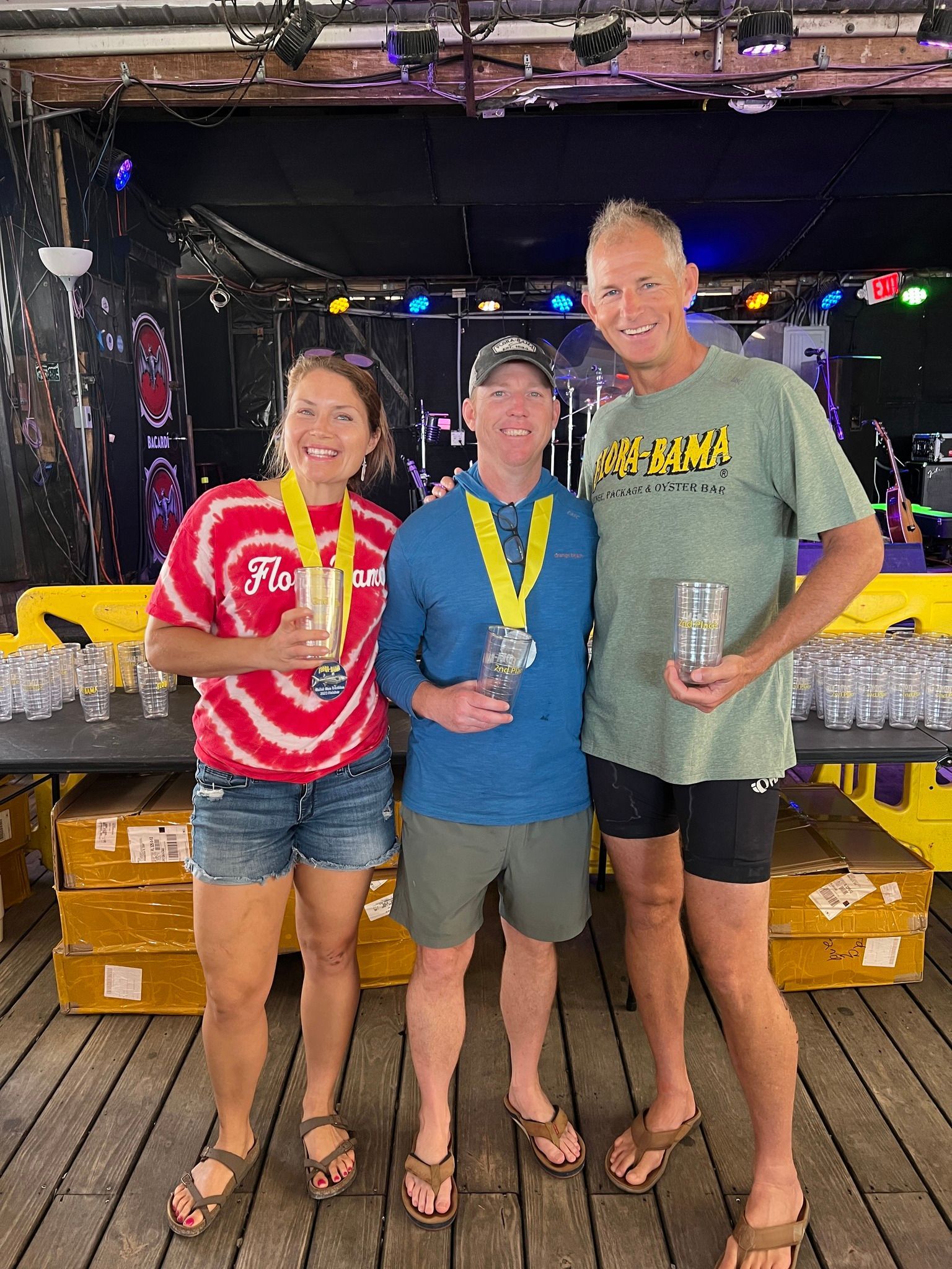 Three people stand on a wooden deck holding awards, with stage lighting and rows of glassware in the background.