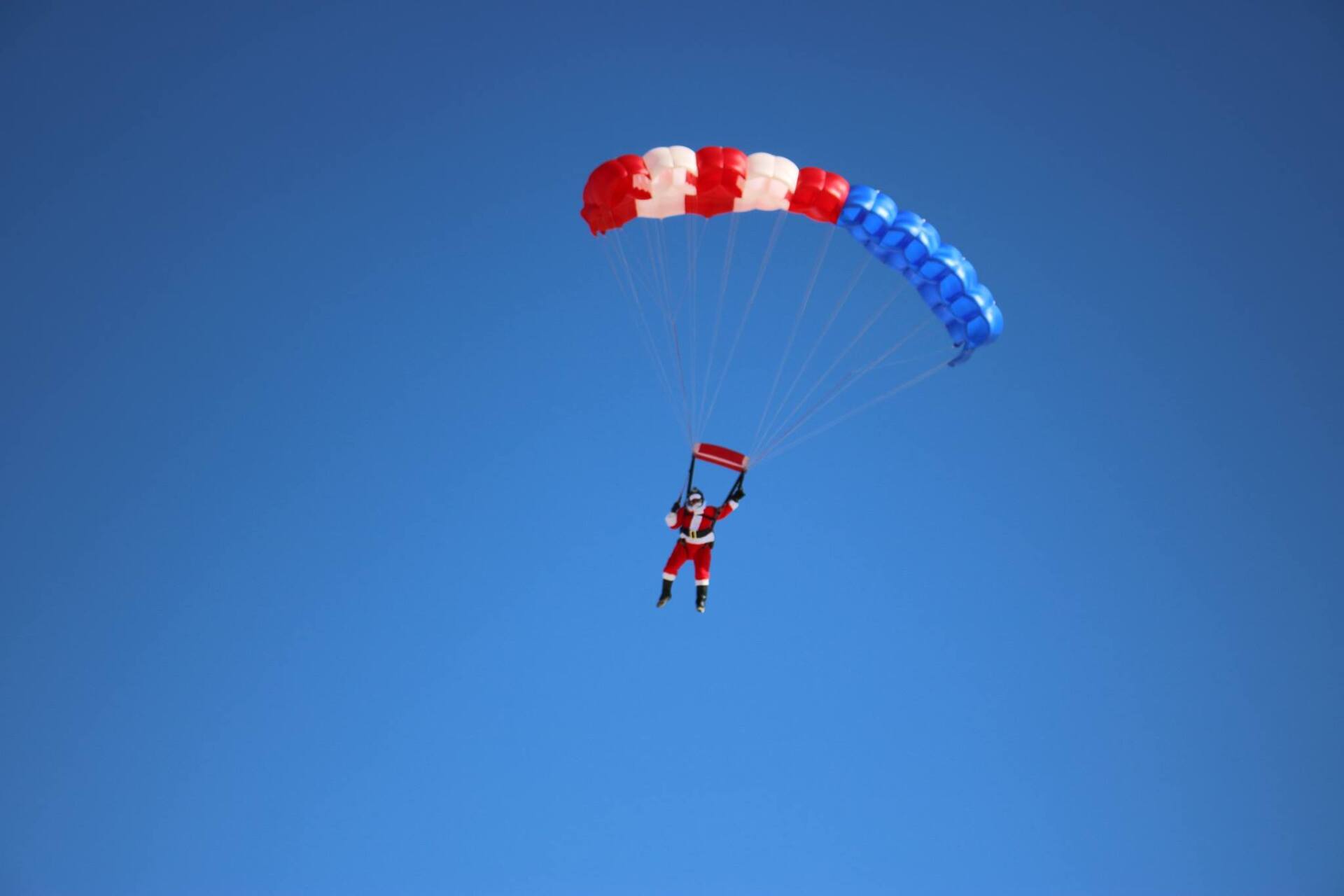 Santa Claus skydiving under a red, white, and blue parachute against a clear blue sky.