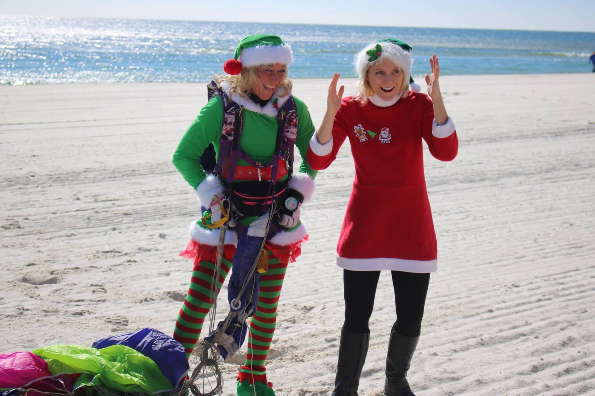 Two people in festive holiday outfits stand on a sandy beach; one wears elf gear with a parachute, the other red attire.