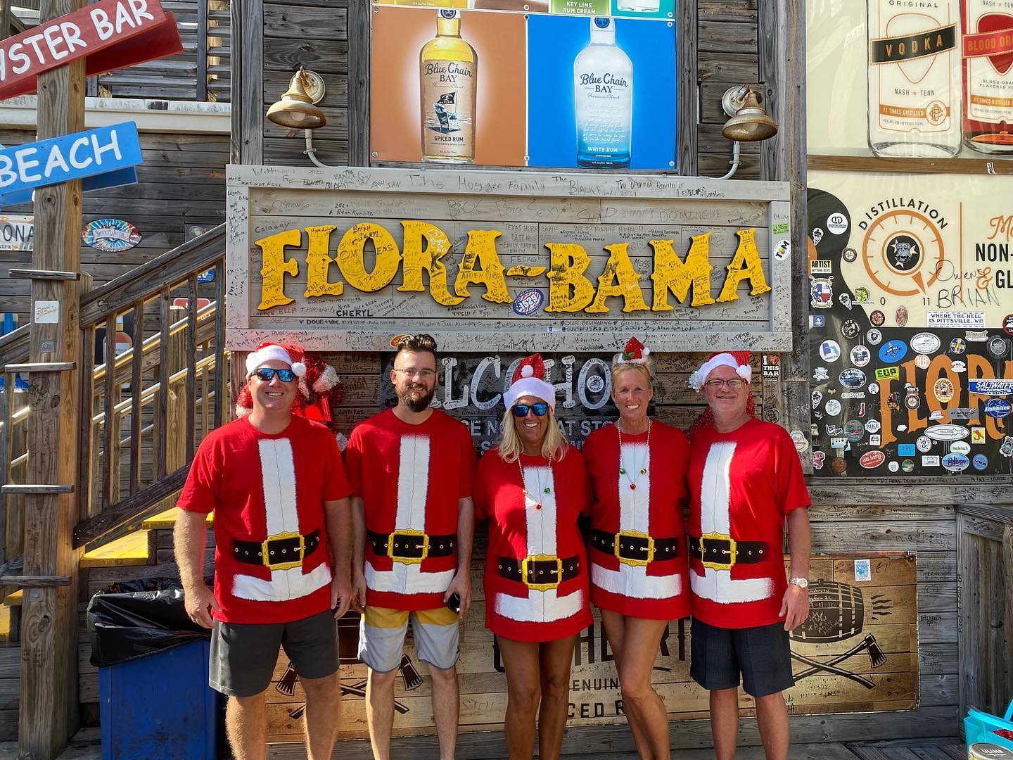 Five people wearing Santa-themed t-shirts pose in front of a Flora-Bama sign at an outdoor bar.