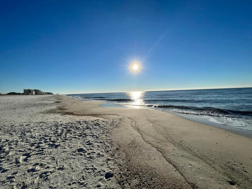 Bright sun shines over a vast, sandy beach with calm waves gently lapping at the shore under a clear blue sky.