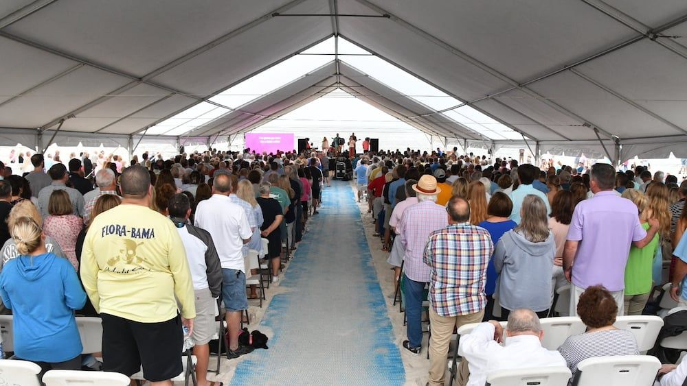 A large crowd stands inside an open-sided white event tent, facing a stage at the far end of a long, blue-carpeted aisle.