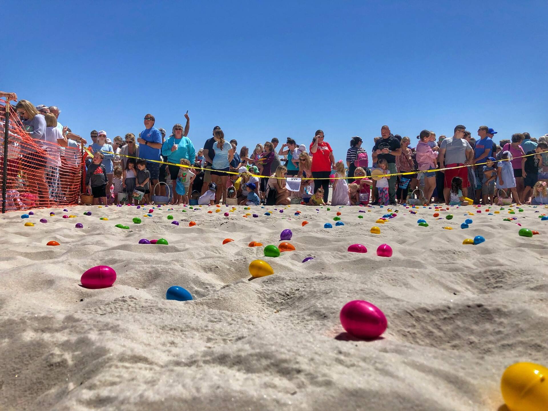 A crowd waits behind a yellow rope at a sunny beach where many colorful plastic Easter eggs are scattered on the sand.