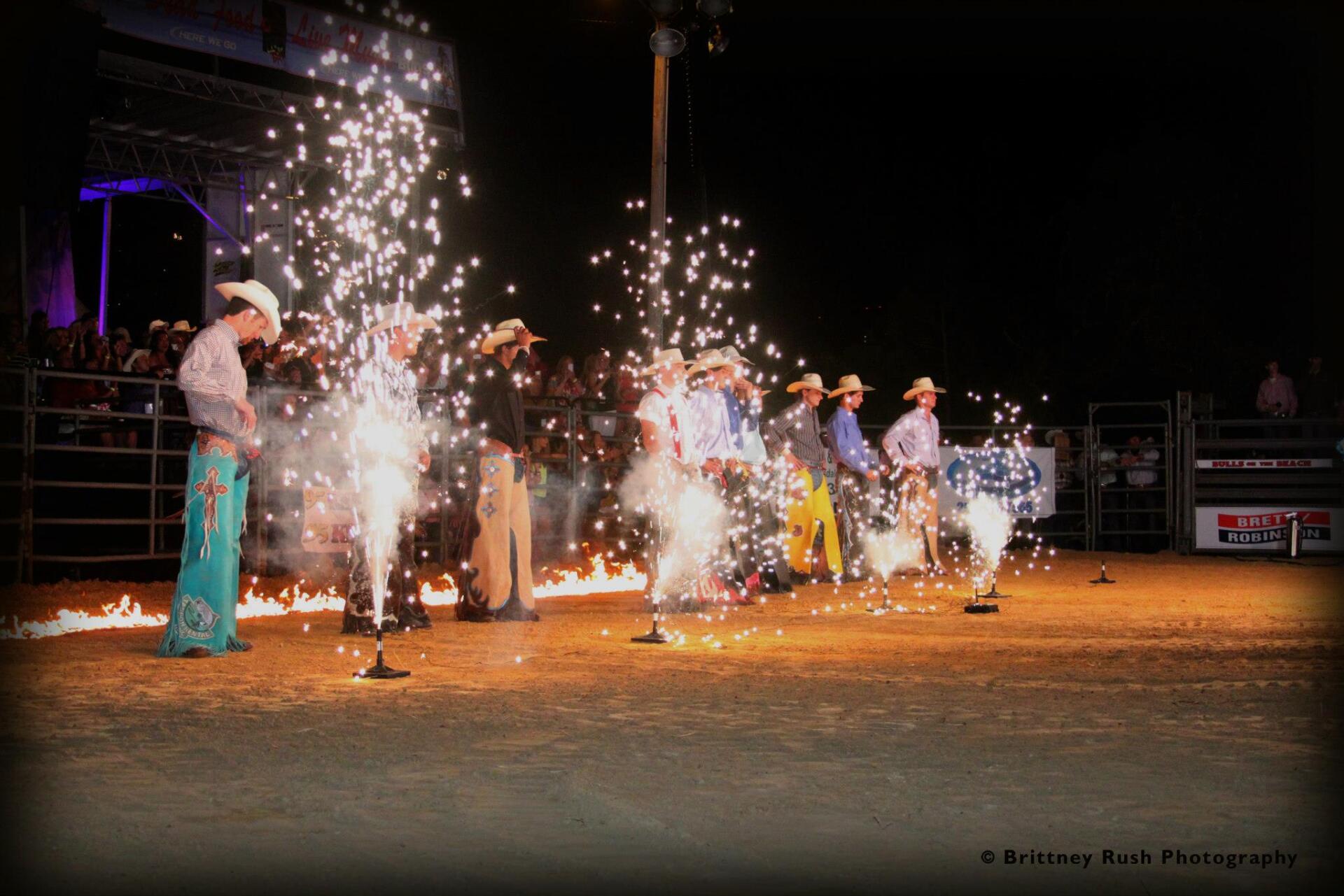 A group of riders in cowboy hats stand in an arena as pyrotechnic fountains spray bright sparks into the air at night.