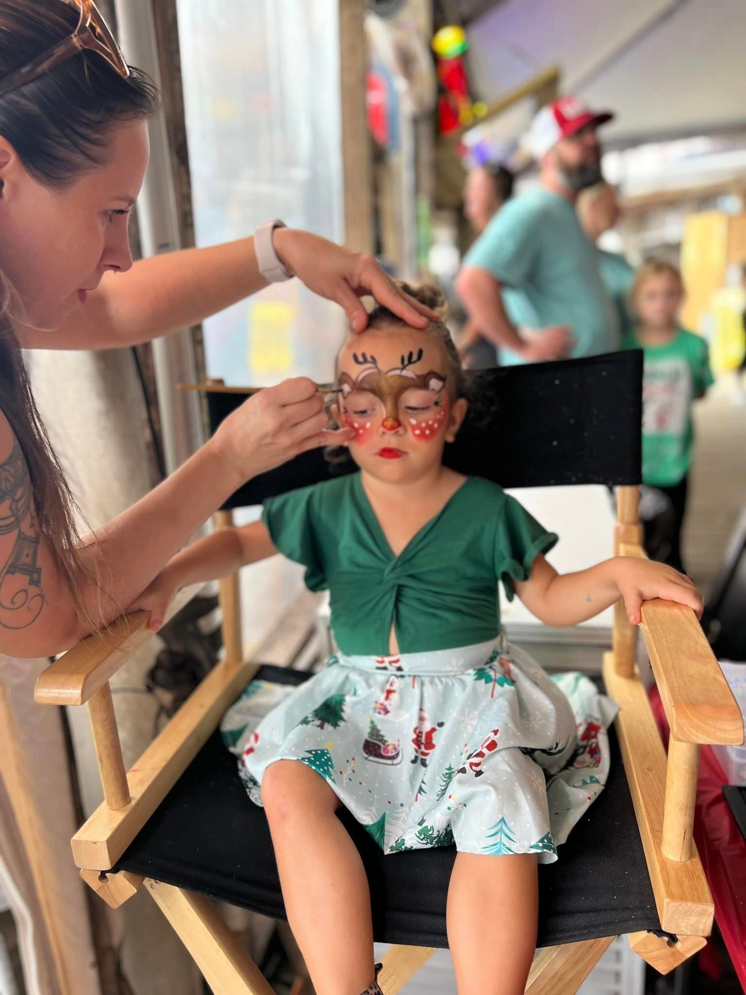 A painter applying festive reindeer makeup to a child sitting in a folding chair while wearing a Christmas-themed outfit.