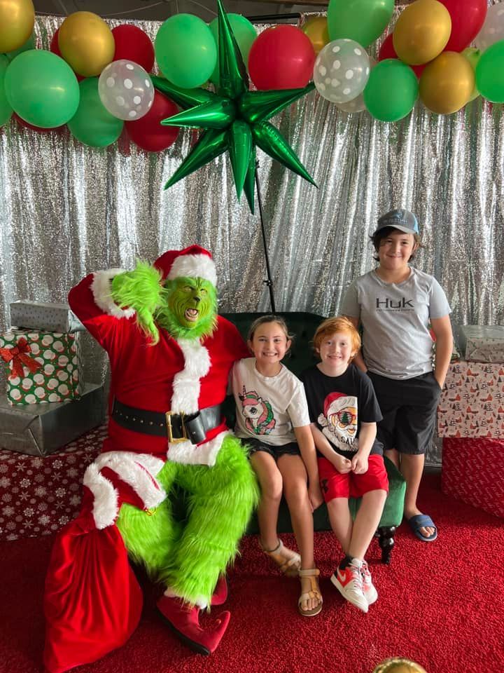 A person dressed as The Grinch sits with three children in front of a festive backdrop with balloons and wrapped gifts.
