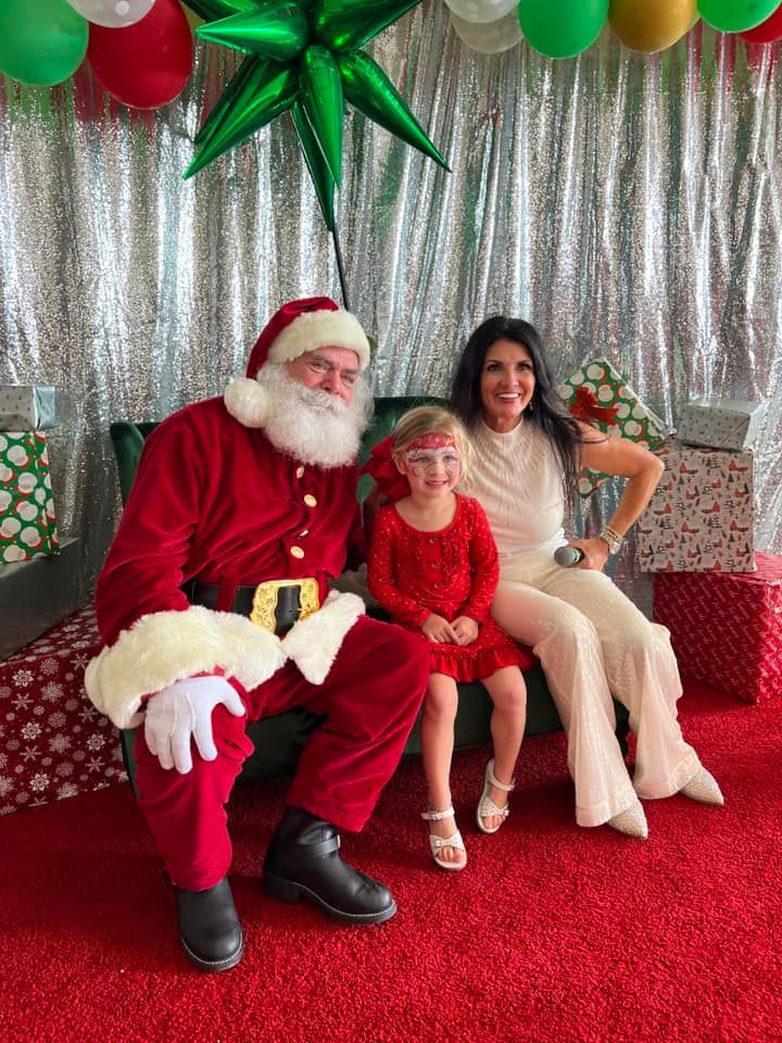 Santa Claus sits with a child and an adult on a bench in front of a silver backdrop with gifts and balloons.