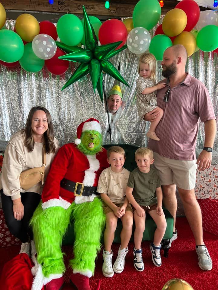 A group posing for a photo with a person dressed as the Grinch in front of a silver tinsel backdrop and festive balloons.