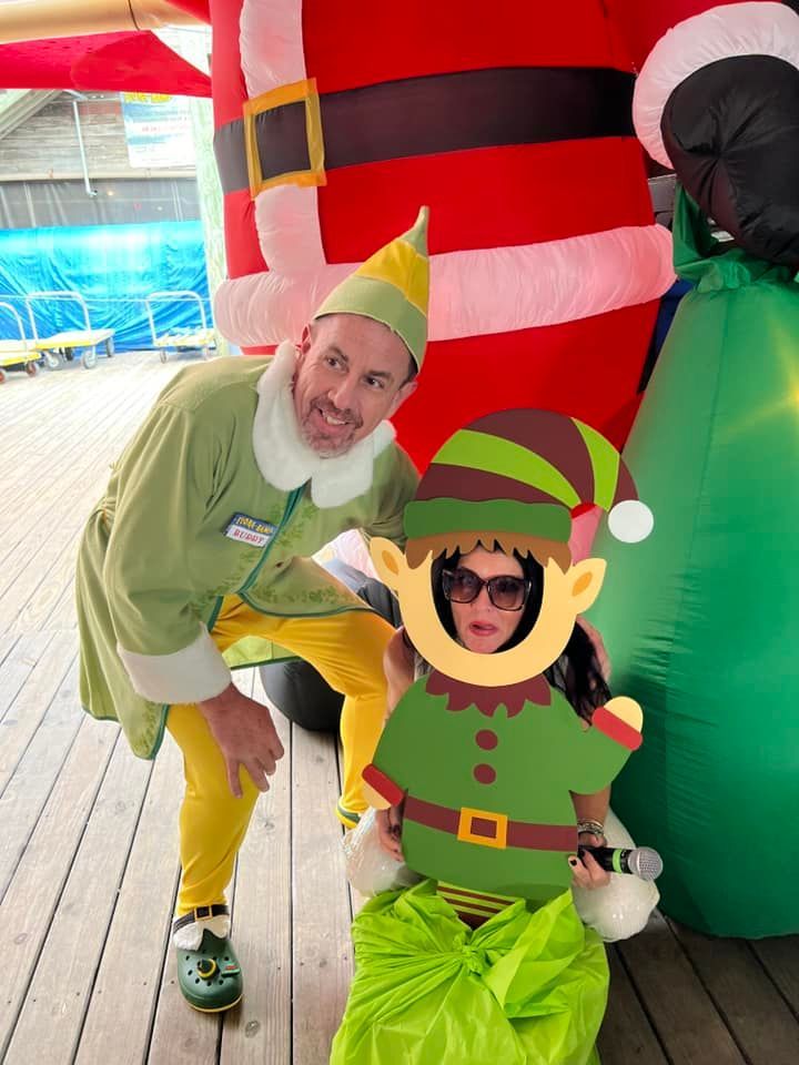 A person in an elf costume stands beside another person posing behind a cardboard elf cutout in front of a giant inflatable.