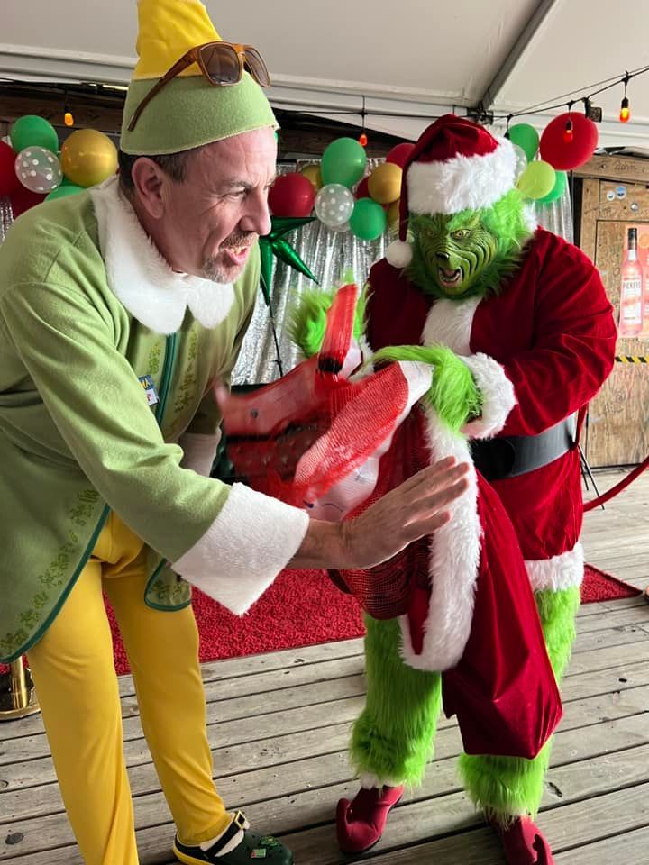 A person dressed as an elf reaches toward a person in a Grinch costume wearing a Santa suit, with balloons in the back.