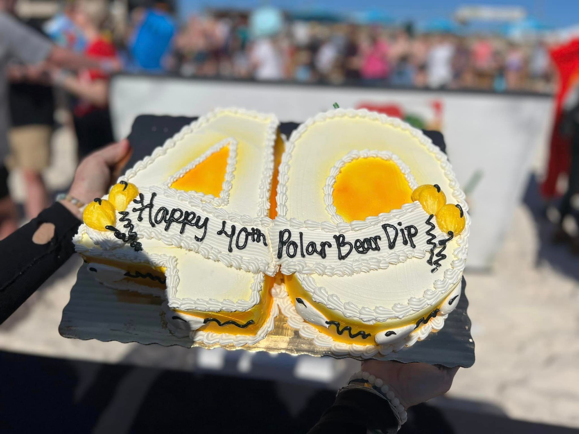 A person holds a number-shaped cake for a 40th Polar Bear Dip at a beach, decorated with yellow frosting and text.