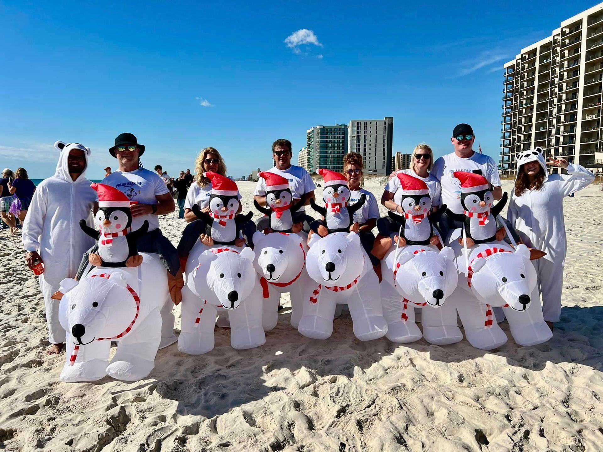 A group in matching Christmas-themed polar bear and penguin costumes on a sunny beach in front of tall resort buildings.