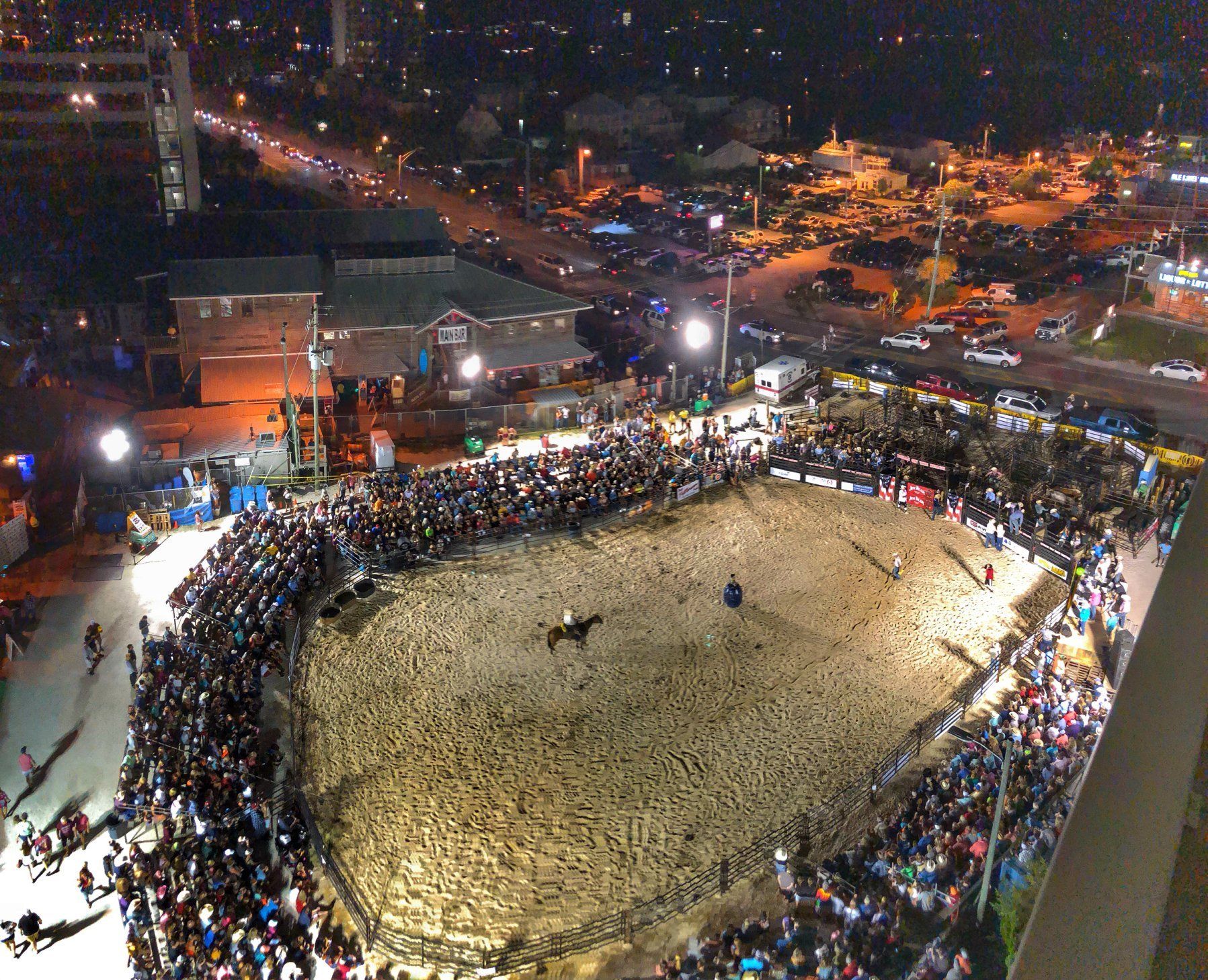 High-angle night view of a crowded arena with a horse and rider in a dirt ring, surrounded by spectators and city lights.