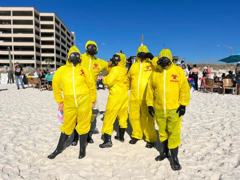 Five people in bright yellow hazmat suits and gas masks stand together on a sunny, sandy beach near a large hotel.