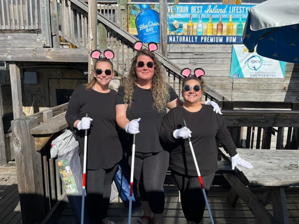 Three people dressed as mice in black outfits, mouse ears, and white gloves, holding canes outdoors on a wooden deck.