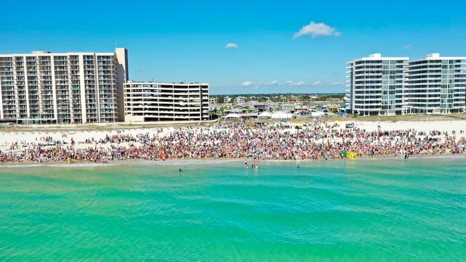 A crowded beach with turquoise water in the foreground and large multi-story apartment buildings in the background.