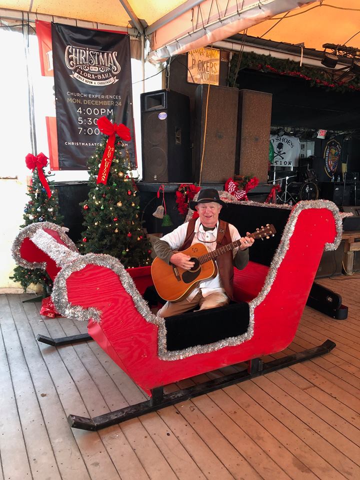 A musician with an acoustic guitar sits inside a decorated red Christmas sleigh in an outdoor pavilion.